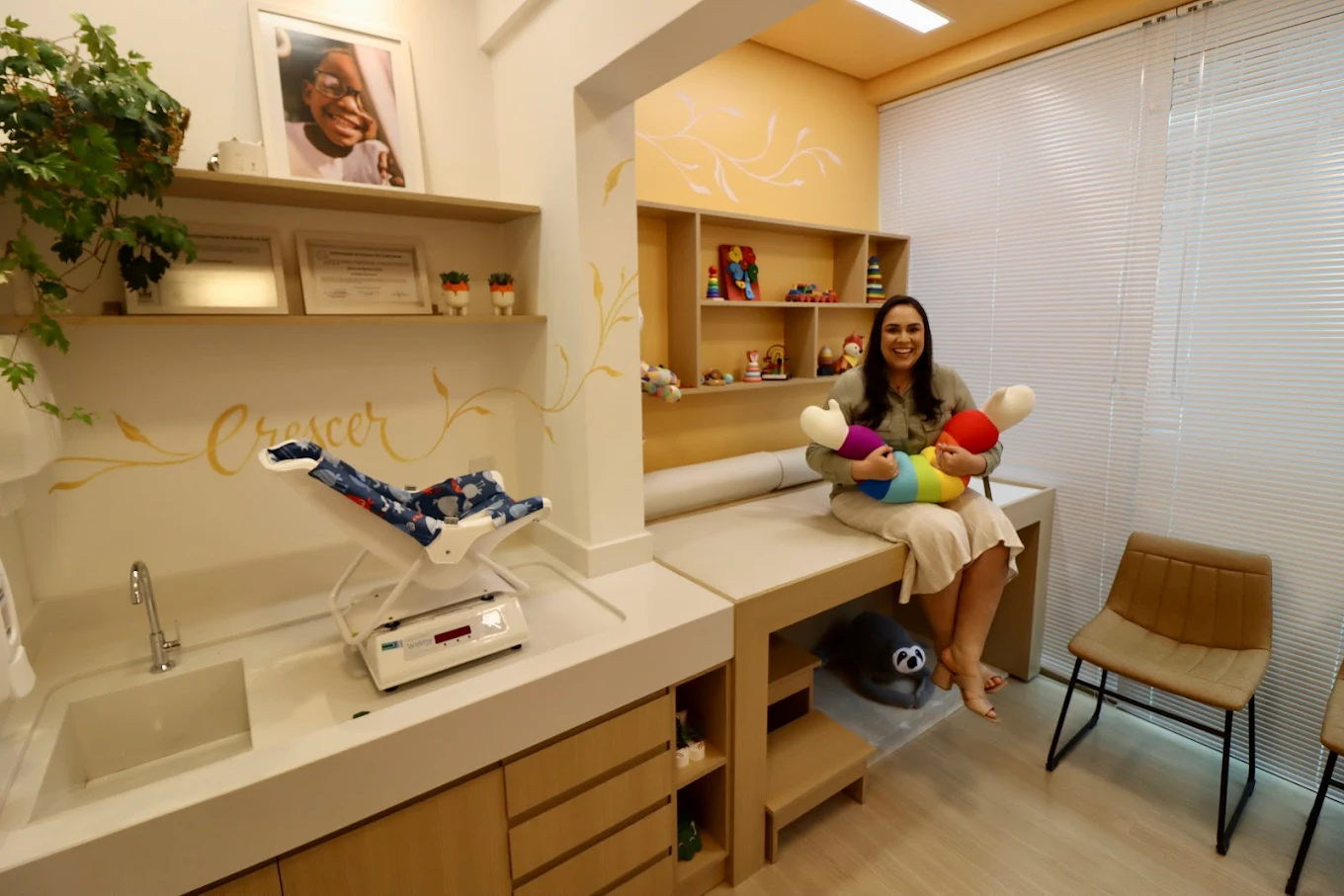 Sala de atendimento com balança pediátrica, prateleiras com brinquedos e a Dra. Clarissa