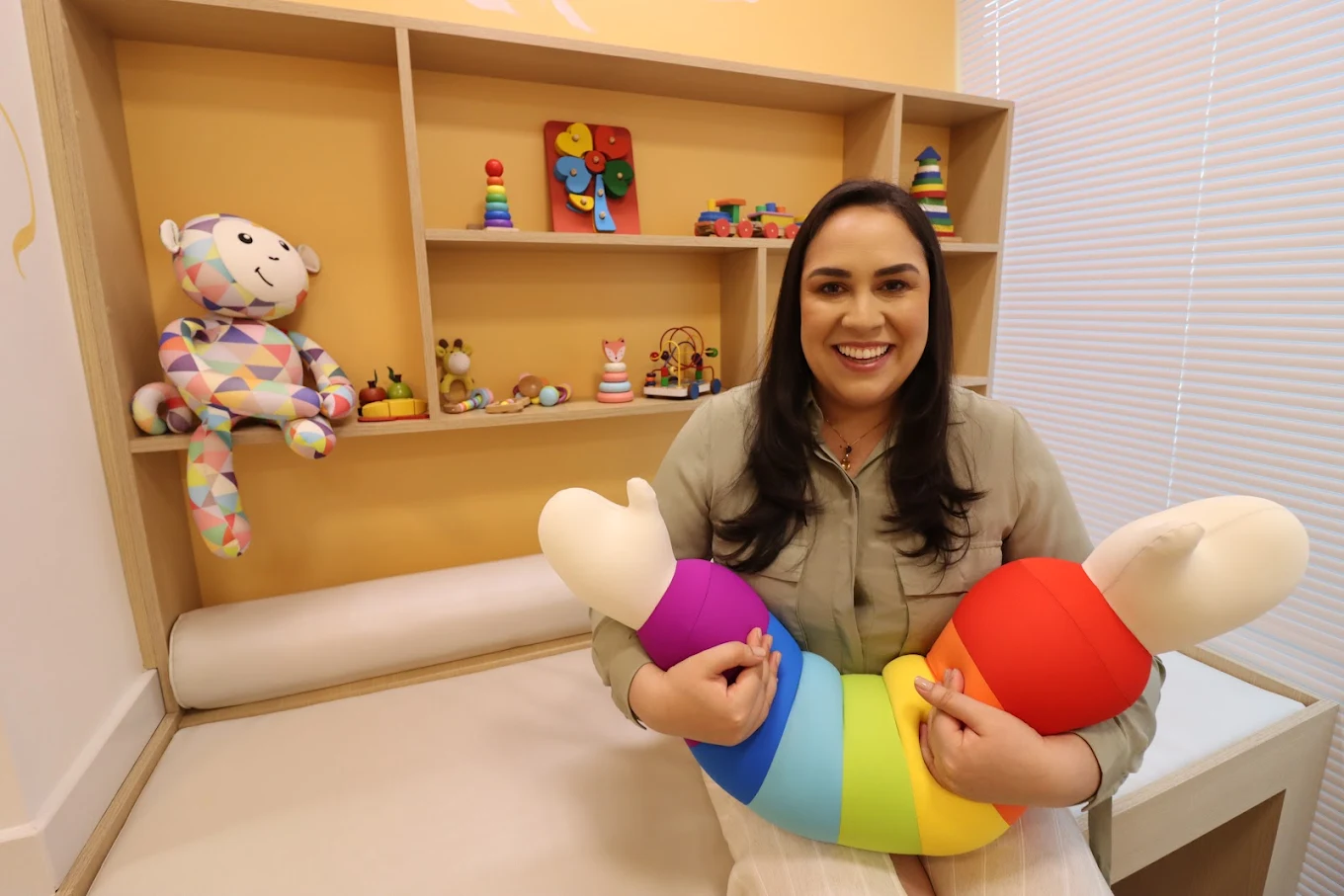 Dra. Clarissa sorrindo na maca pediátrica com brinquedos ao fundo